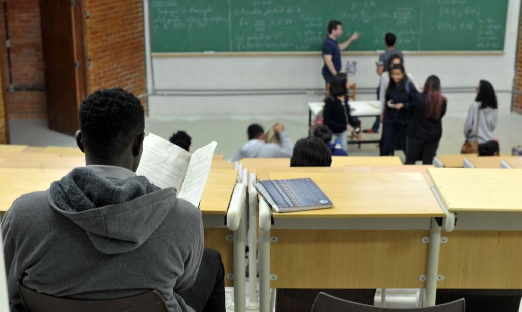 Sala de aula universitária. No plano de fundo, estudantes fazem contas na lousa. No primeiro plano, um estudante negro lê um livro.