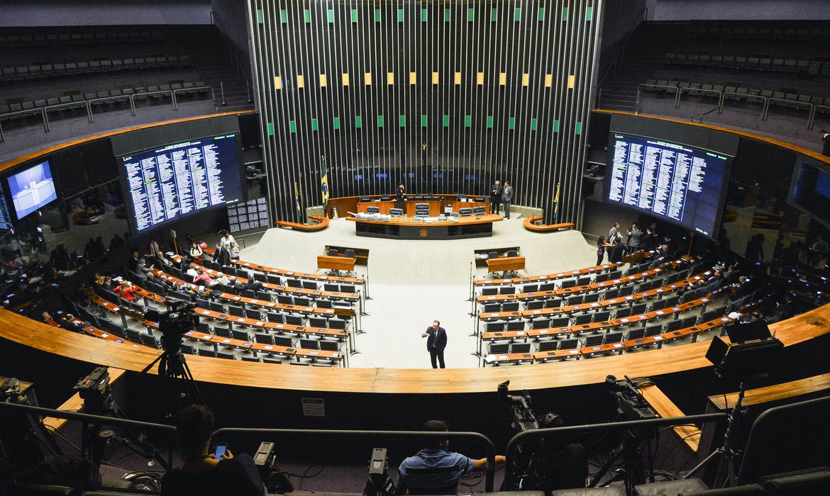 Plenário da Câmara dos Deputados, em Brasília. Sala oval com cadeiras ordenadas em circulo e um telão ao fundo.